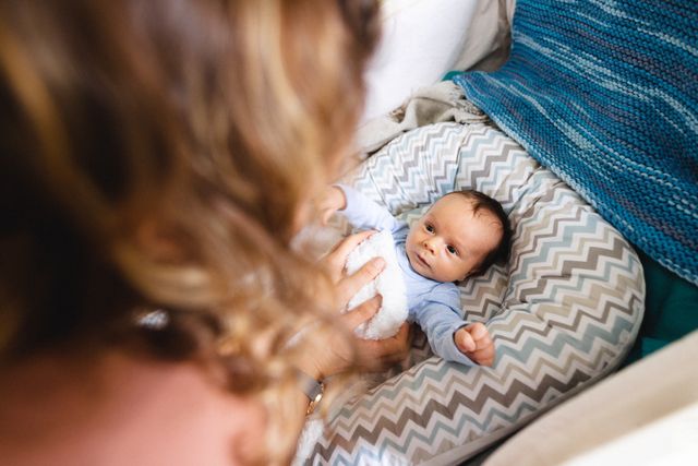 High angle view of a mother holding her newborn baby in a cozy home environment. The baby is lying on a patterned pillow, looking up at the mother. This image can be used for parenting blogs, family-oriented advertisements, articles on motherhood, and baby care products.