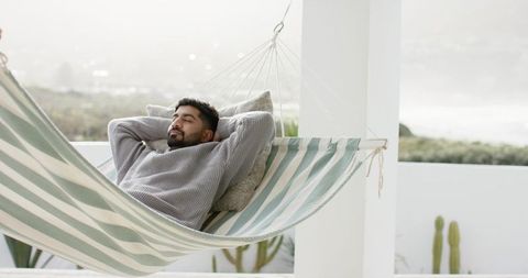Indian man reclining in striped hammock on coastal balcony relaxing in cozy sweater