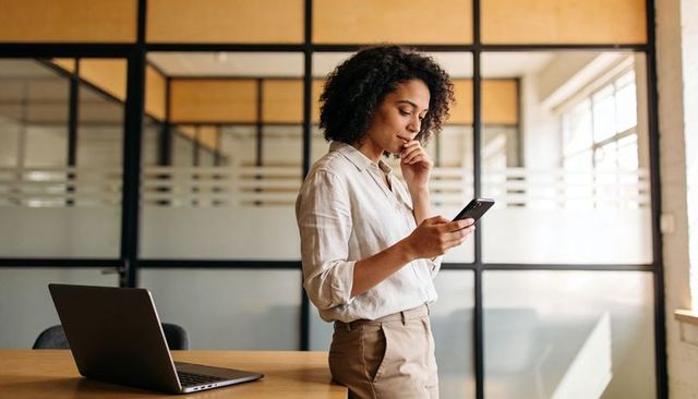 Thoughtful businesswoman checking smartphone in sunlit modern office with laptop on desk