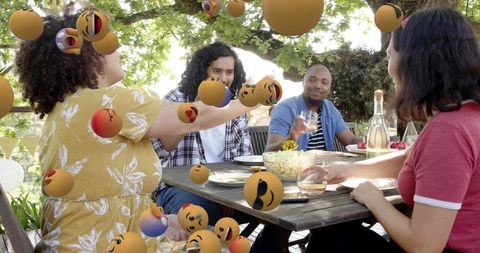 Friends chatting and clinking glasses at backyard table with floating emoji spheres