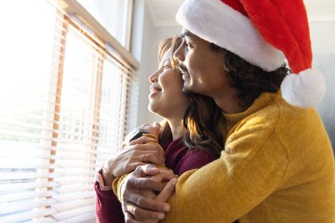 Diverse couple hugging by sunlit window wearing santa hat and cozy sweaters holiday morning