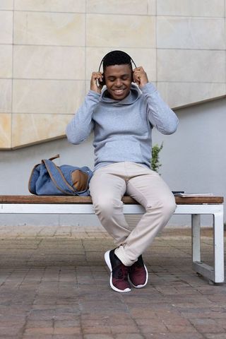 African american young man listening to music on bench with backpack in urban plaza casual outfit