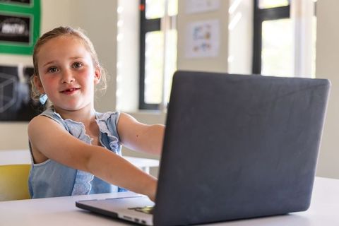 Elementary school girl using laptop and reaching across desk in bright classroom