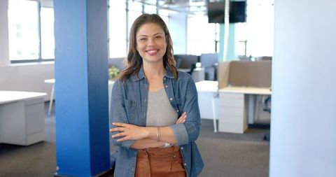 Confident professional woman standing with arms crossed smiling in modern open-plan office