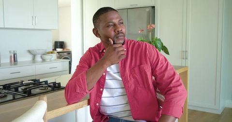 African american man sitting at kitchen island gazing thoughtfully wearing red shirt