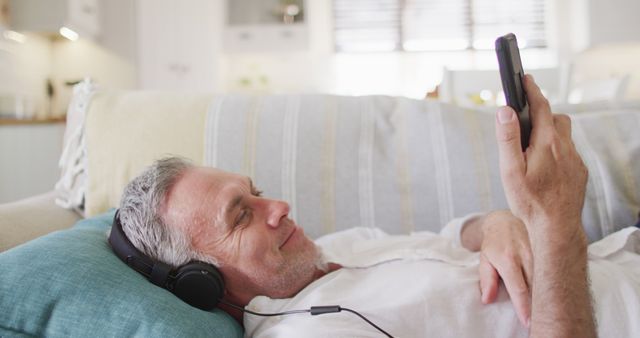 Senior man wearing headphones and using smartphone while lying on couch. Can be used for concepts related to leisure time, elderly technology use, relaxation, home comfort, and audio enjoyment.