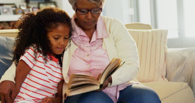 An African American senior woman is sharing a book with a young girl, her granddaughter, creating a warm, educational moment. Their close bond is evident as they enjoy a story together, fostering a love for reading.