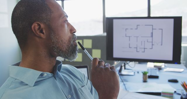 An architect is concentrating on a building floor plan displayed on a computer screen at his office. This is useful for illustrating concepts related to architecture, designing, professional workplaces, and intellectual pursuits.