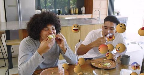 Young men eating avocado sandwiches at kitchen table with playful emoji overlays