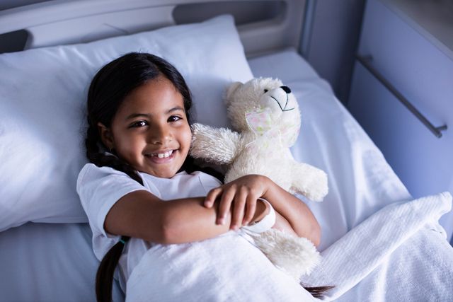 Young girl lying in hospital bed, hugging teddy bear and smiling. Ideal for healthcare, pediatric care, recovery, and medical treatment themes. Can be used in brochures, websites, and advertisements related to children's hospitals, patient care, and emotional support.