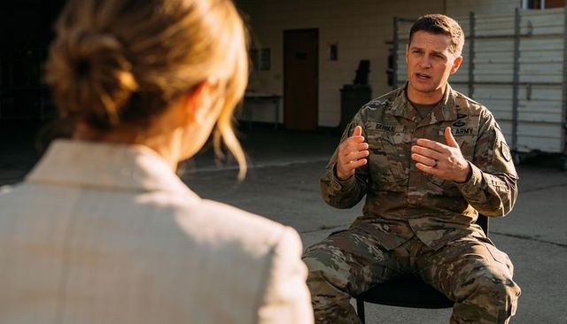 Sunlit army officer speaking to interviewer in hangar wearing camouflage uniform, gesturing