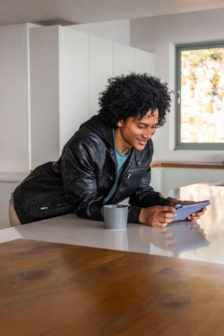 African american man leaning on kitchen island using tablet and smiling in modern home