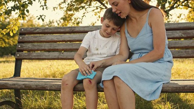 Sunlit caregiver tending scraped knee, comforting boy while sitting on wooden park bench