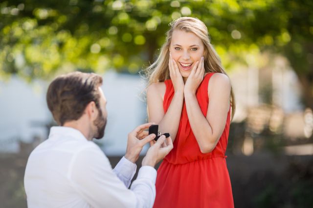 Man proposing to woman with a ring in a park. Woman is surprised and happy, wearing a red dress. Ideal for use in content about engagements, romantic moments, relationships, and marriage proposals.