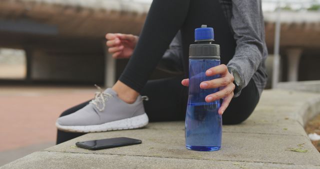 Woman sitting on a concrete step holding a blue water bottle with a smartphone beside her after a workout. She is wearing gray sneakers and dark leggings with a gray long-sleeve shirt. Ideal for content focusing on fitness, hydration, healthy living, exercising outdoors, or workout routines.