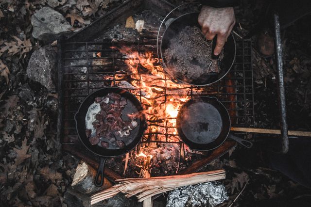 Close-up of person cooking meat and food in cast iron skillets over a campfire. Flames are visible along with firewood and rocks surrounding the fire. Ideal for content related to camping, outdoor adventures, wilderness cooking, hiking, and rustic living.