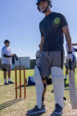 Young cricket batter gearing up on sunny practice pitch with teammates preparing gear