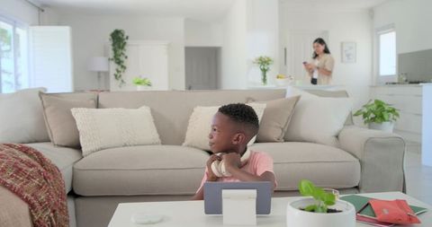 Child sitting at coffee table using tablet with headphones in sunlit modern living room