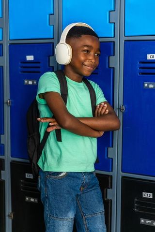 Confident schoolboy standing at lockers wearing headphones and backpack, smiling