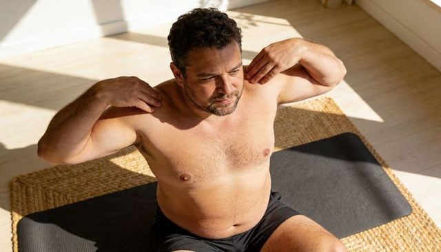 Middle-aged man stretching shirtless on yoga mat in sunlit home workout