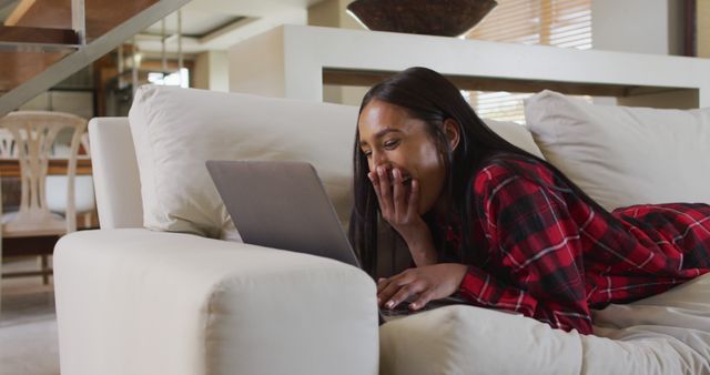 Young woman in plaid shirt lying on sofa, engaged with laptop and smiling. Ideal for concepts around remote work, online communication, leisure activities, modern lifestyle, technology use, and comfortable living.