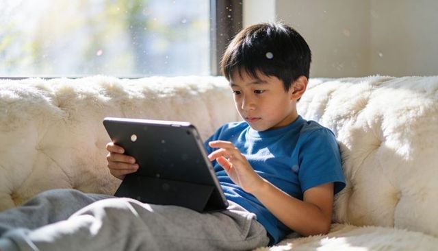 Sunlit boy using tablet on plush couch focusing on remote learning and quiet screen time