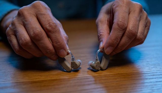 Senior hands holding behind-ear hearing aids on wooden table closeup for hearing care