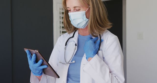Female doctor wearing gloves and face mask, holding a tablet in a hospital room. Ideal for health-related articles, medical publications, healthcare apps, promotional materials for hospitals or clinics, and educational resources about patient care and use of technology in healthcare.