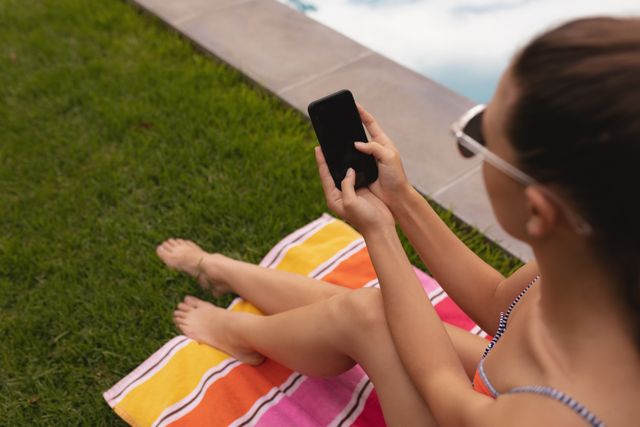 This image shows a Caucasian woman in swimwear sitting on a colorful towel by the poolside in a backyard. She is using a mobile phone, suggesting a moment of relaxation and leisure. This image can be used for themes related to summer activities, outdoor relaxation, technology use in leisure time, and backyard pool settings. Ideal for advertisements, blog posts, or social media content about summer fun, poolside relaxation, or mobile technology.