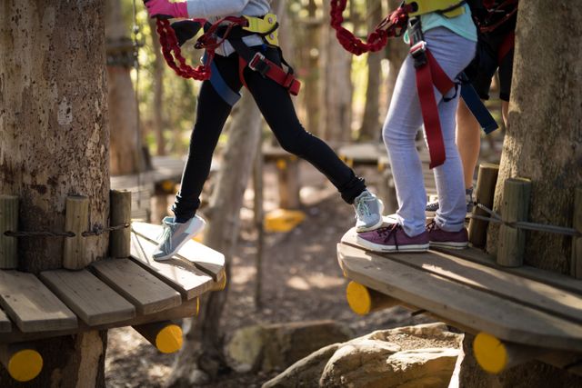 Children crossing a zip line in an adventure park, wearing safety harnesses and balancing on wooden platforms. Ideal for use in promotions for outdoor activities, adventure parks, team-building exercises, and children's physical fitness programs.