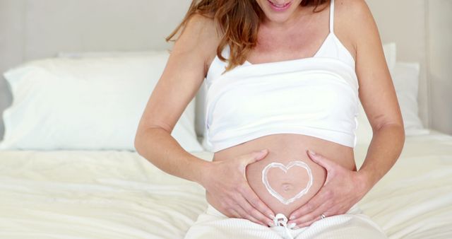 Seeing happy pregnant woman drawing heart shape on her belly with cream is heartwarming. Use for maternity blogs, pregnancy healthcare articles, skincare product advertisements, mother-to-be health guides, prenatal class invites, childbirth preparation tutorials, expecting announcements.