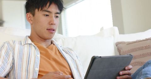 Asian man relaxing on sofa using tablet for streaming and browsing in bright living room