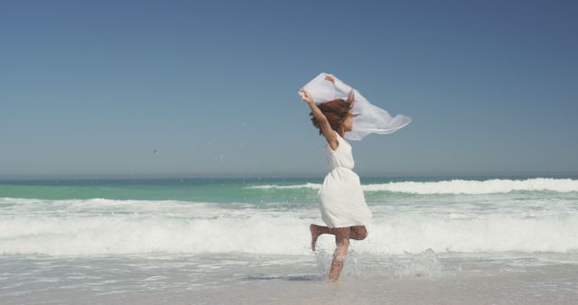 Woman in white dress enjoying sunny day at beach, holding white scarf while jumping among waves. Perfect for travel and tourism promotions, summer-themed content, advertisements, health and wellness articles, and carefree living advertisements.