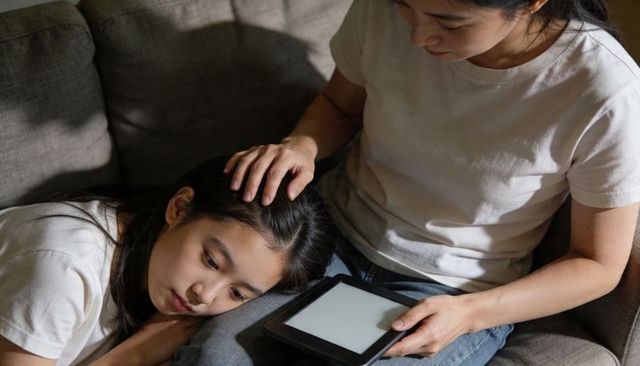Asian mother comforting teen daughter on sofa holding tablet, intimate bonding and care