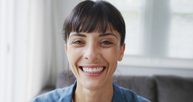 Smiling brunette woman shown indoors with natural light illuminates her face creating a warm and welcoming ambiance. Ideal for use in health and wellness campaigns, promotional materials for positive messaging, and websites focused on happiness and lifestyle themes.