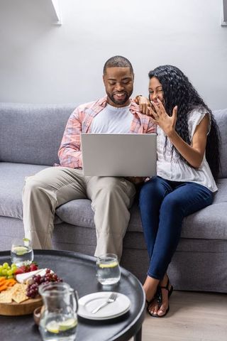 Engaged couple celebrating surprise ring reveal while browsing laptop on cozy gray sofa