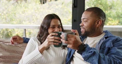 Sharing cozy morning coffee, laughing multiracial couple toasting mugs on couch