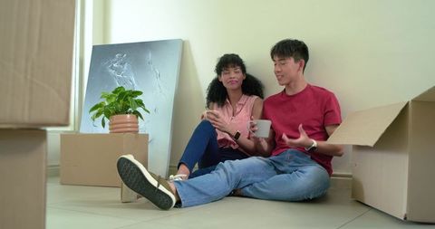 Diverse couple relaxing and unpacking in new apartment with moving boxes and potted plant