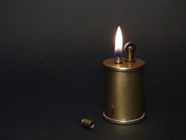 Image depicts a vintage brass lighter with a lit flame against a dark background. Cap is removed and placed beside the lighter. Ideal for use in articles or designs related to retro or vintage collections, smoking accessories, antique collectibles, or old-timey aesthetics.