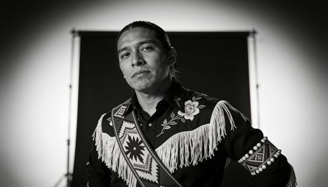 Stoic portrait of man wearing fringed embroidered regalia and beaded sash, monochrome