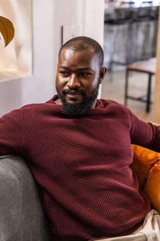 African american man relaxing on modern sofa in coworking lounge wearing maroon sweater