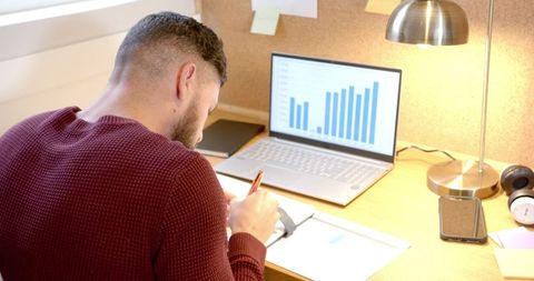 Male Using Laptop And Writing Notes In Home Office Environment
