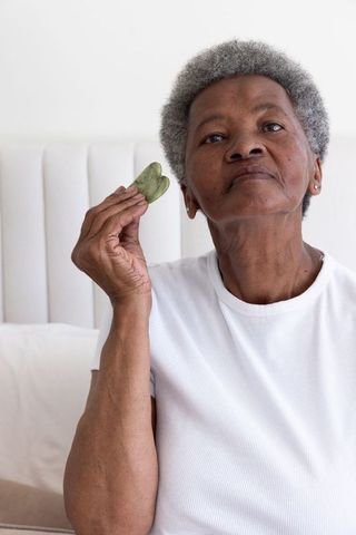 Senior african woman practicing gua sha with heart-shaped jade for facial skincare and wellness