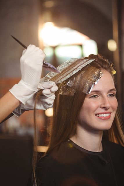 Hairdresser applying hair dye to client's hair using foil and brush at a salon. The client is smiling and appears relaxed. This image is ideal for use in beauty and hair care advertisements, salon promotions, and articles about hair treatments and professional hairstyling services.