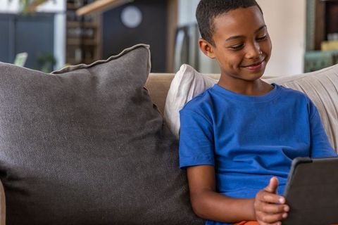 Preteen african american boy smiling while using tablet on cozy living room sofa