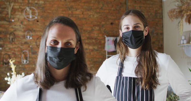 Female bakery staff in casual and striped aprons, wearing face masks, standing in front of a stylish brick wall backdrop. Suitable for content on food service, COVID-19 safety measures, small businesses during the pandemic, or features on female entrepreneurship in the bakery industry.