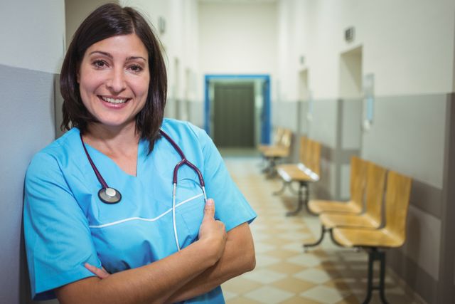 A female surgeon in blue scrubs, smiling and standing with arms crossed in a hospital corridor. Suitable for use in medical-related articles, healthcare promotions, hospital advertisements, and educational materials on careers in the medical field.