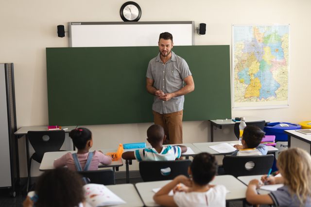Male teacher standing in front of a blackboard, instructing a group of diverse elementary school students seated at desks. Classroom includes educational materials and a map on the wall. Ideal for use in educational content, school brochures, and articles about teaching and learning environments.