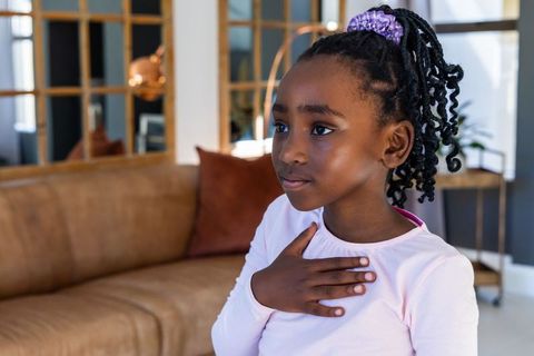 Young girl standing in living room holding hand to chest wearing purple scrunchie