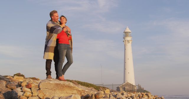 Couple embracing warmly while standing on rocky beach with a tall lighthouse in the background at sunset. Ideal for travel promotions, romantic getaways, outdoor adventures, and advertisements highlighting intimate, scenic moments.